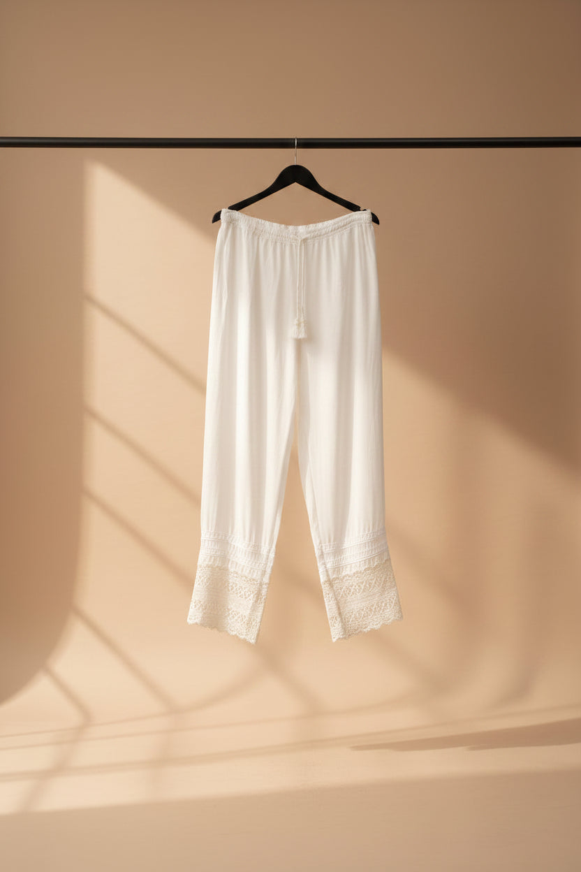 White cropped pants with lace trim at the hem, hanging on a wooden hanger outdoors against a backdrop of potted plants and tree branches.
