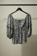 A black and white patterned off-the-shoulder blouse with puff sleeves, hanging on a wooden hanger against a dark red background.