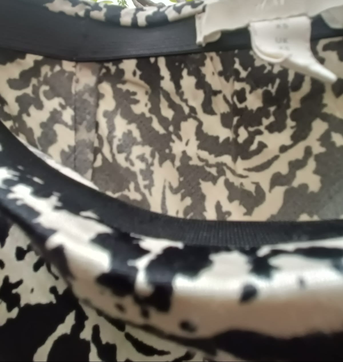 Close-up of a black and beige abstract patterned garment, showing the inner neckline with black trim and a white sewn-in size tag.
