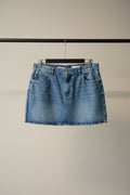 A light blue denim mini skirt with a frayed hem, hanging on a wooden hanger against a solid red background.