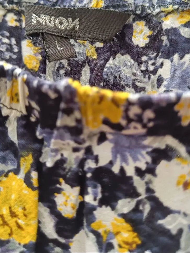 Close-up of a floral-patterned garment with a visible NUOM brand label. The fabric has a navy, white, and yellow abstract floral print, and the photo is taken outdoors with blurred tree trunks and leaves in the background.