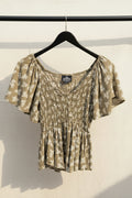 An olive green and white floral print blouse with puff sleeves and a smocked bodice, hanging on a wooden hanger against a dark red background.
