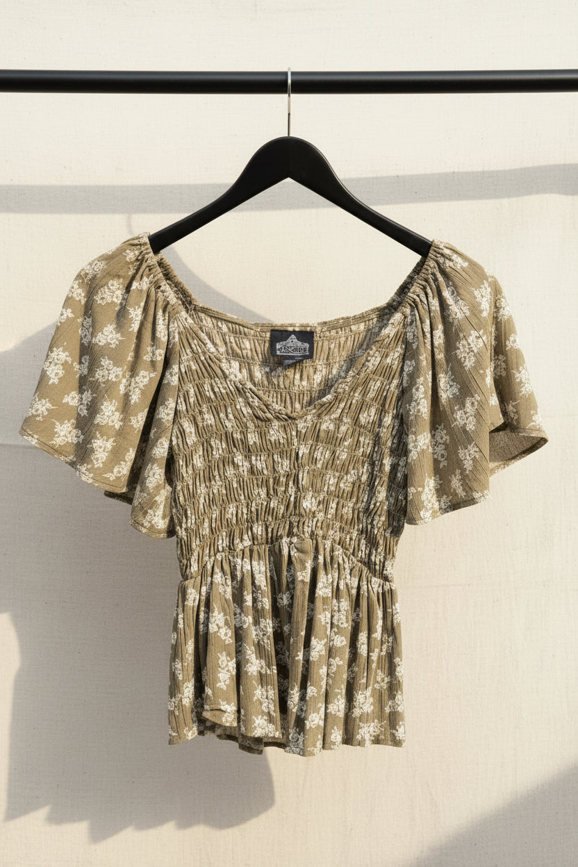 An olive green and white floral print blouse with puff sleeves and a smocked bodice, hanging on a wooden hanger against a dark red background.