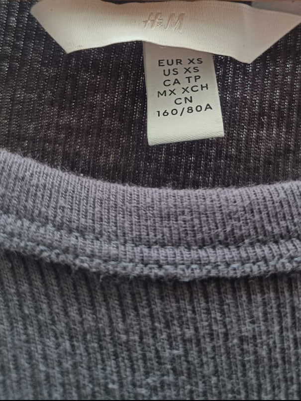 Close-up of a dark gray ribbed knit crewneck sweater hanging on a wooden hanger, showing the inner neck label with size information and the H&M logo.