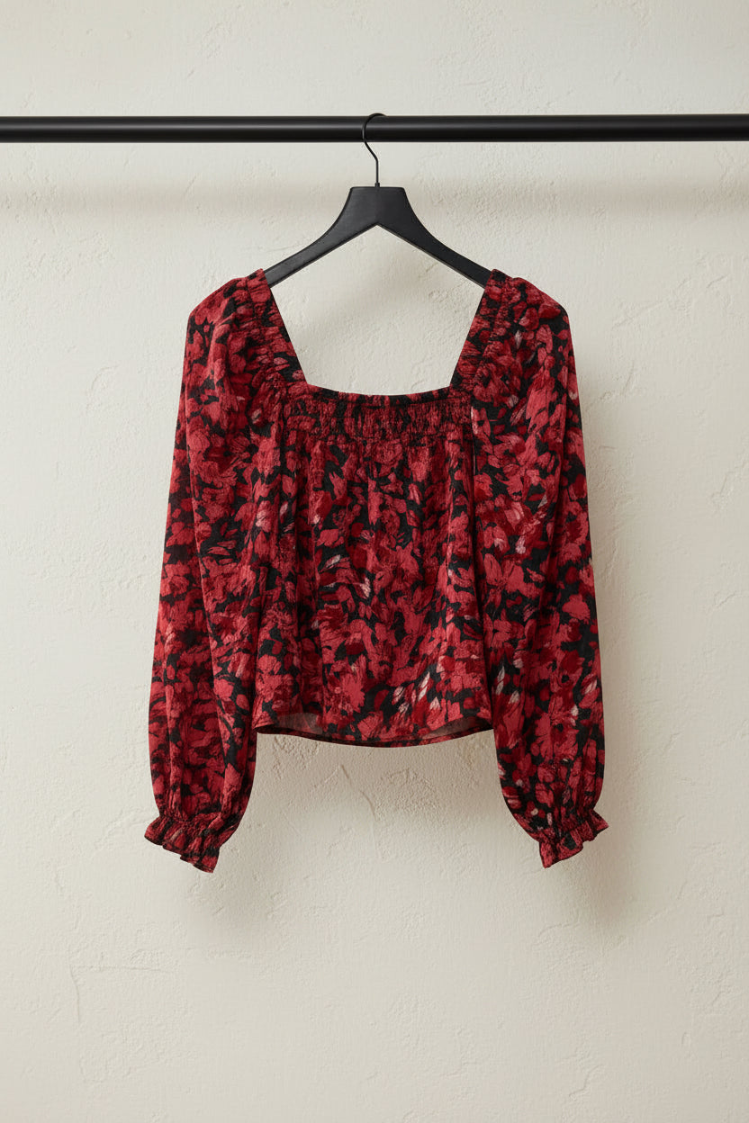 A long-sleeve cropped blouse with a red and black abstract floral pattern, laid flat on a white fabric background.