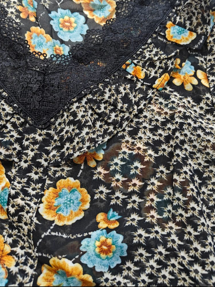 A close-up of a black sheer floral blouse with a dense white star-like print, large yellow and blue flowers, and a black lace V-neckline.